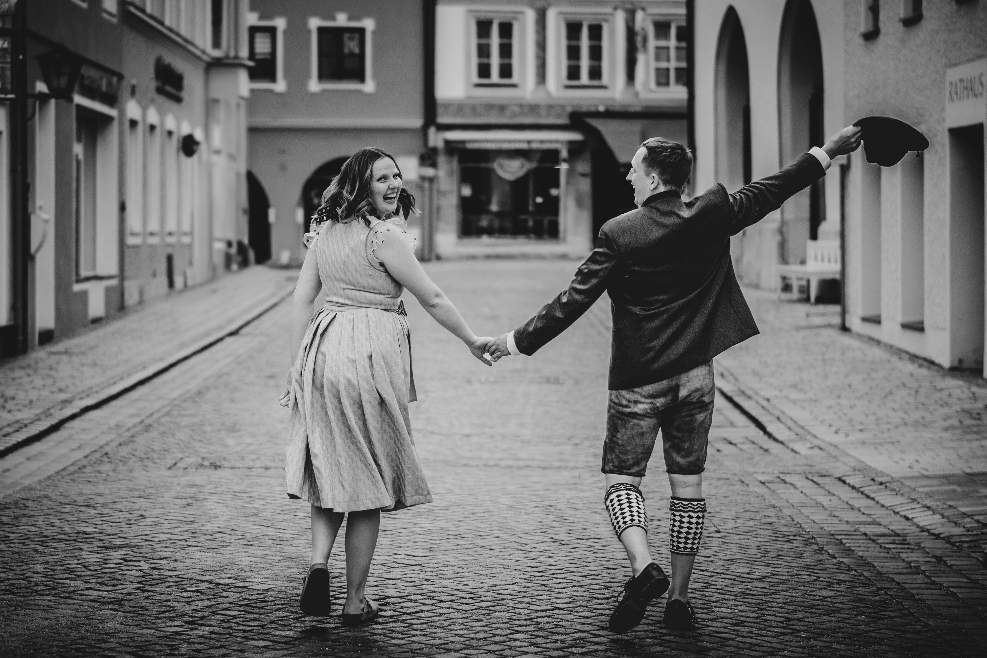 Paar in Tracht geht lachend durch eine Altstadtgasse, der Mann schwenkt seinen Hut